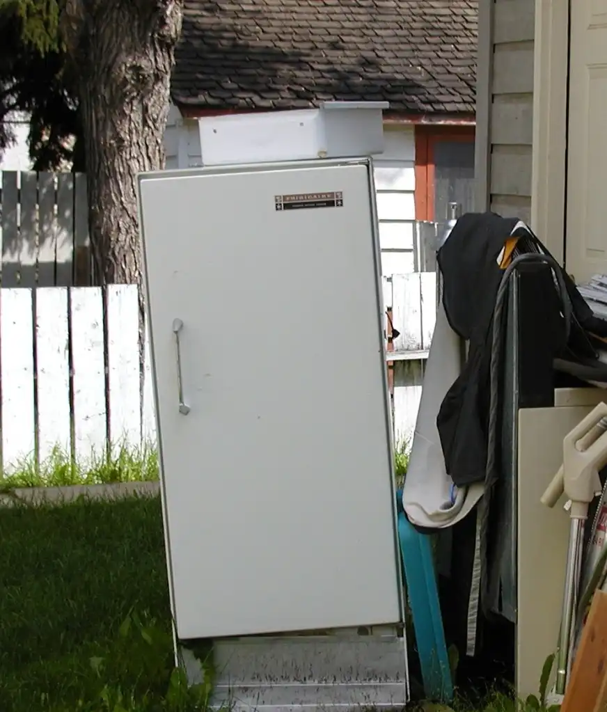 A vintage white refrigerator leaning against a wall in an outdoor setting.